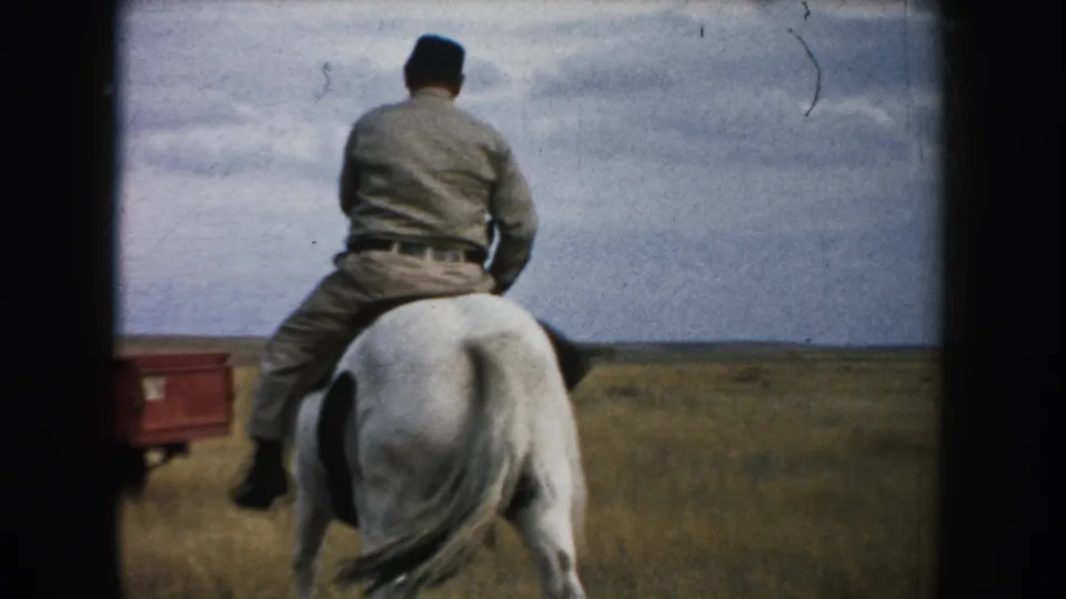 Rear view of an adult rider on a brown-and-white horse in South Dakota grassland, with a red pickup visible at left.