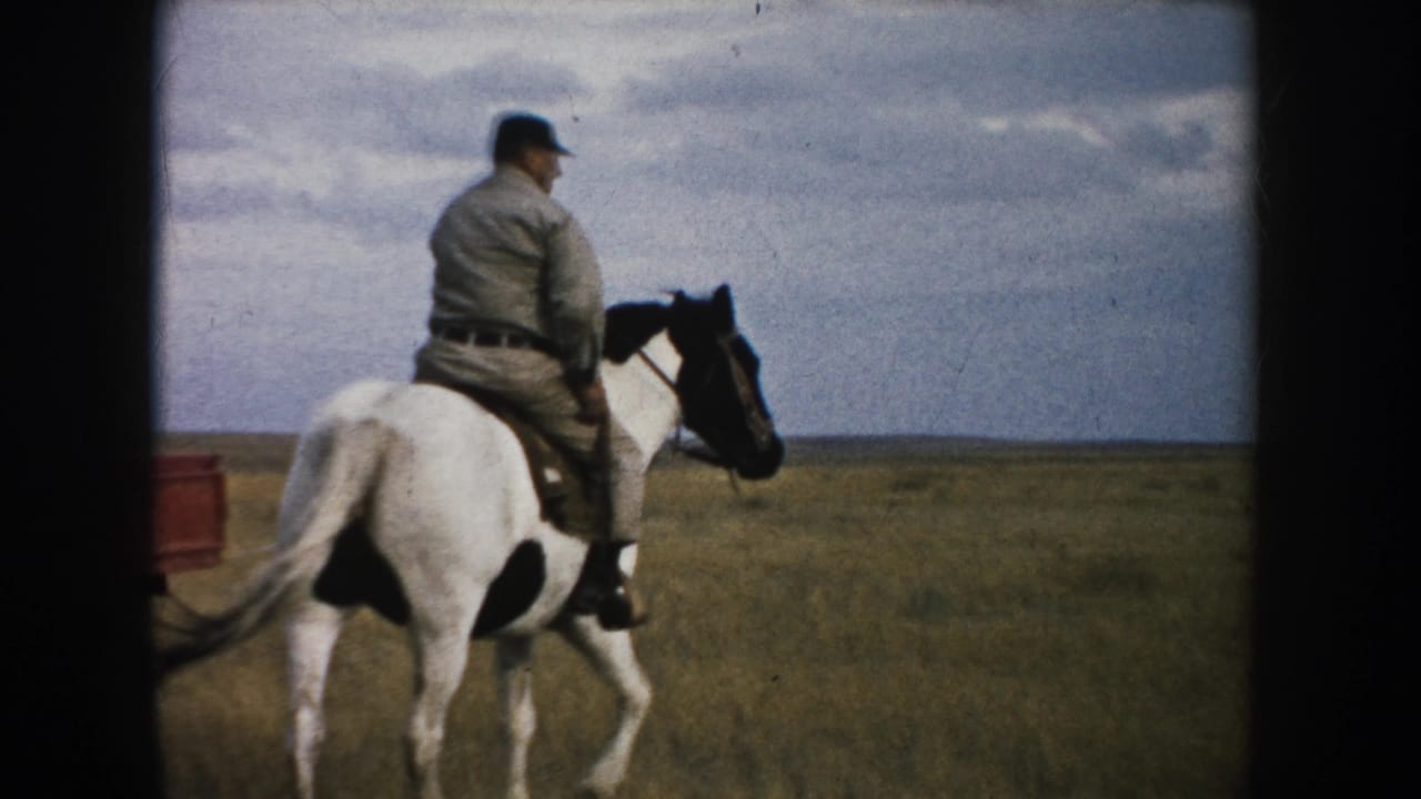 Adult rider on a brown-and-white horse moves through South Dakota grassland, with part of a red pickup at the left edge.