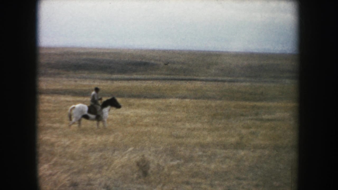 A rider on a pinto horse crosses an open grassy prairie under a cloudy sky.