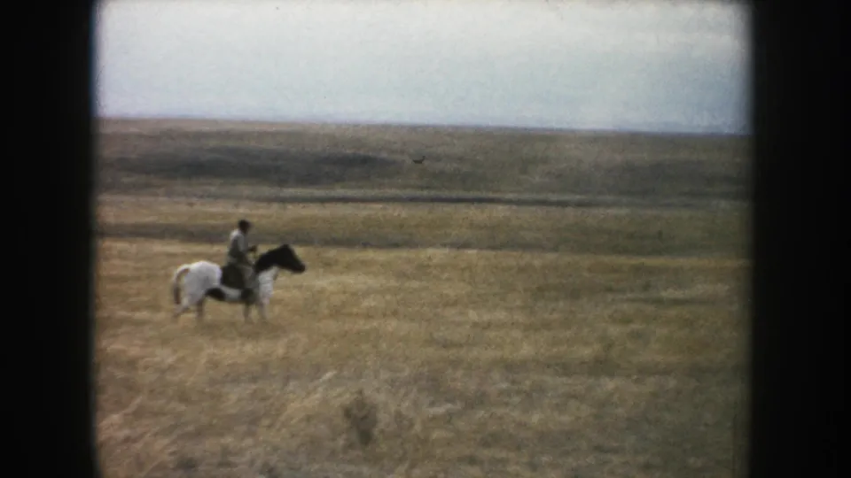 1953 South Dakota home-movie frame showing a man riding a pinto horse across open prairie grass.