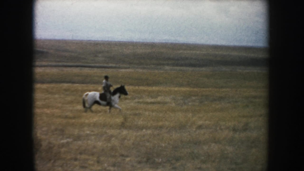 1953 South Dakota frame with a man riding a pinto horse through open prairie under an overcast sky.