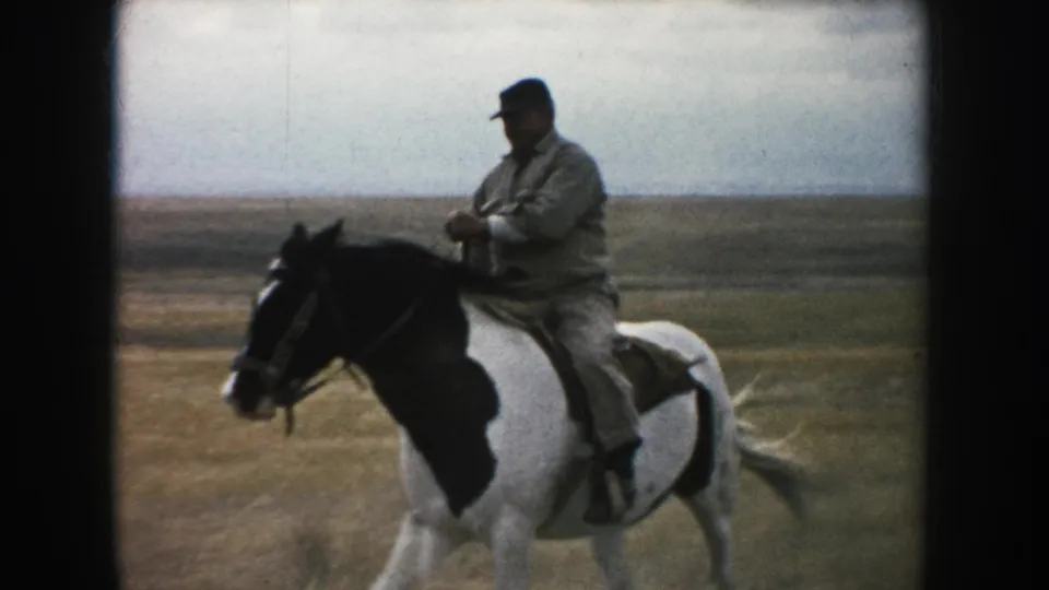 1953 South Dakota frame showing a closer side view of the man riding a pinto horse in open grassland.
