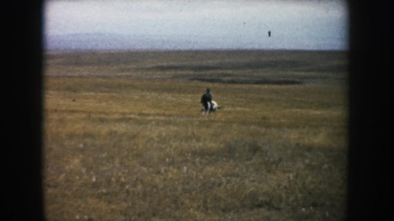 Wide 8mm view of a lone rider and horse moving through South Dakota prairie grass with a low, open horizon.