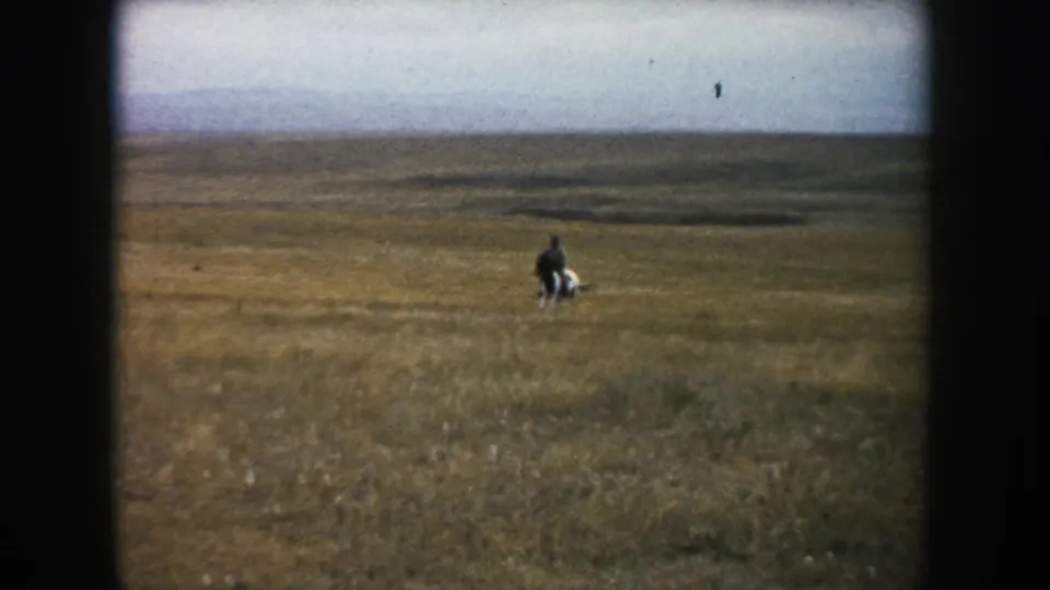 Wide 8mm view of a lone rider and horse moving through South Dakota prairie grass with a low, open horizon.