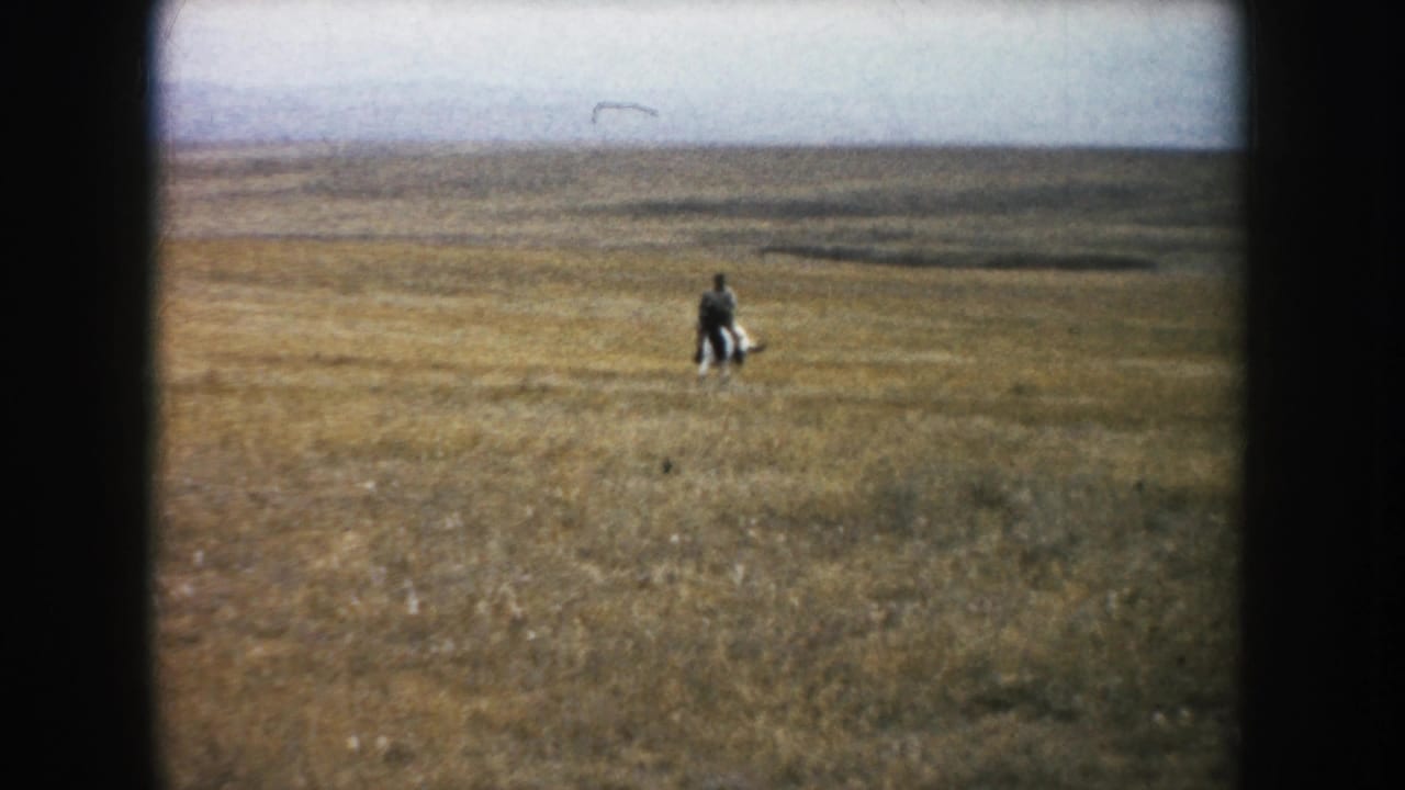 Distant South Dakota prairie frame showing an adult rider on horseback moving through open grassland beneath a pale sky.