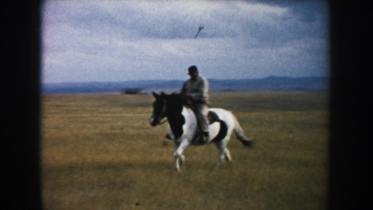 The pinto horse carries an adult rider across muted prairie grass, with a cloudy sky and distant hills behind them.