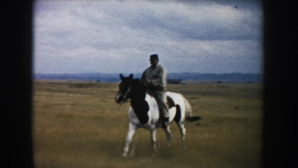 An adult rider on a black-and-white pinto horse approaches through dry South Dakota prairie in vintage 8mm home-movie footage.