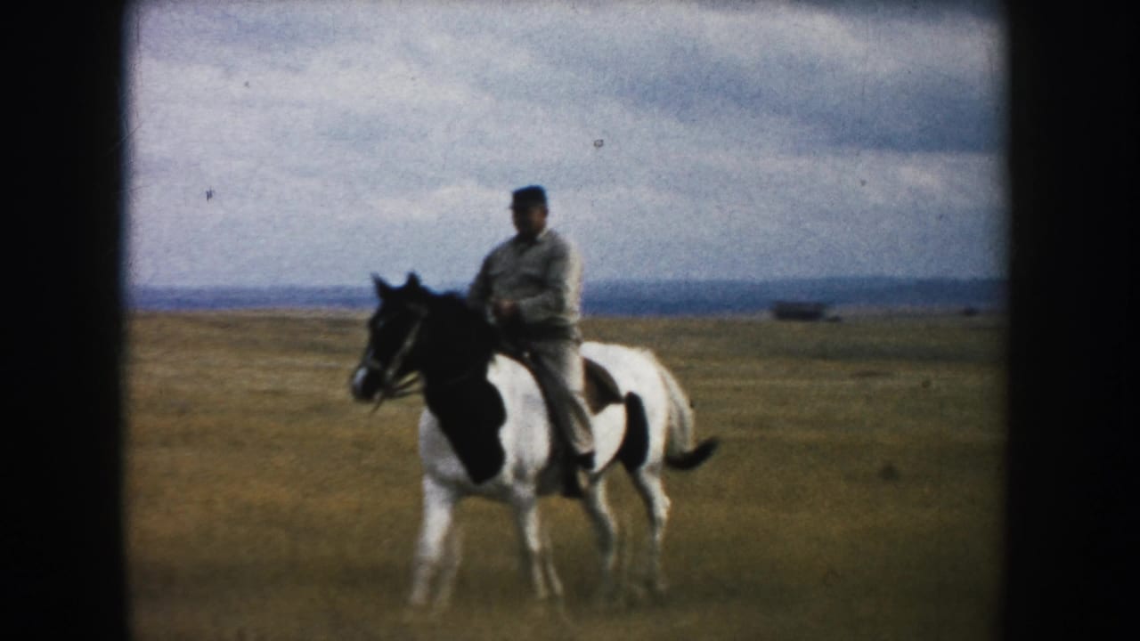 Man rides a black-and-white pinto horse across open prairie under a cloudy sky.