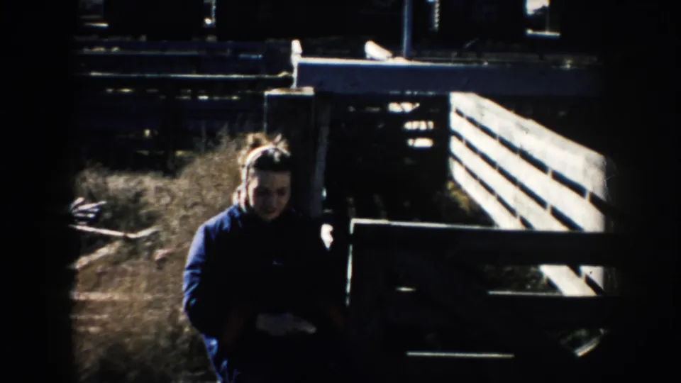 1953 South Dakota home-movie frame showing a woman near wooden stockyard pens, with strong shadows and farmyard fencing around her.