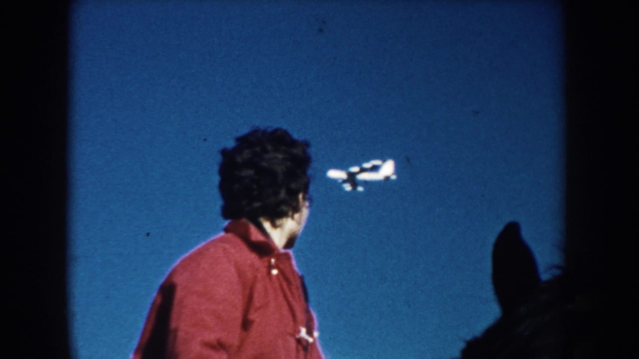 1953 South Dakota home-movie frame of a person in a red jacket looking up as a small airplane crosses the clear blue sky.