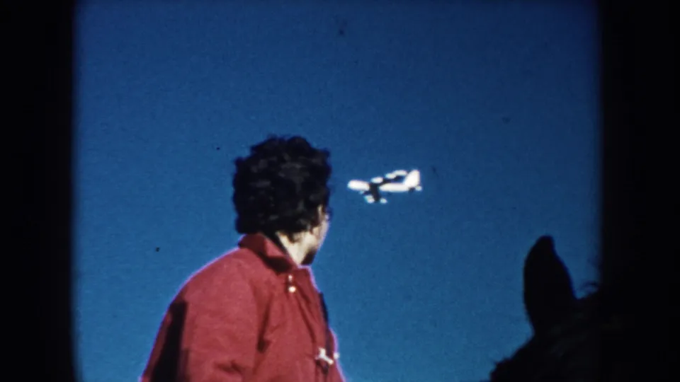 1953 South Dakota home-movie frame of a person in a red jacket looking up as a small airplane crosses the clear blue sky.