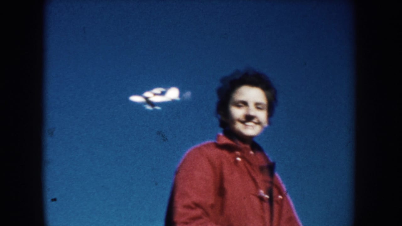 1953 South Dakota 8mm frame showing a smiling person in a red jacket with a small airplane visible in the blue sky behind them.