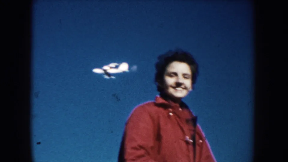 1953 South Dakota 8mm frame showing a smiling person in a red jacket with a small airplane visible in the blue sky behind them.