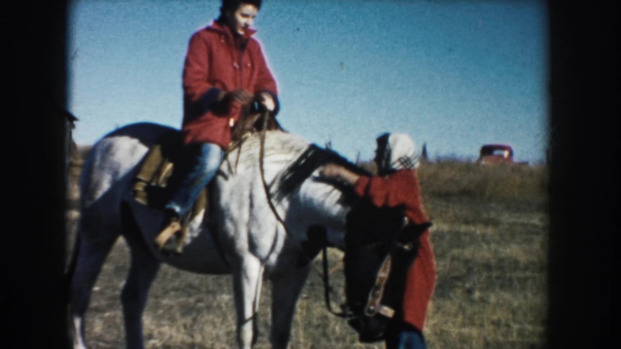 In a 1953 South Dakota field, a man in a red jacket sits on a white horse as a woman in a red coat stands near the horse's head.