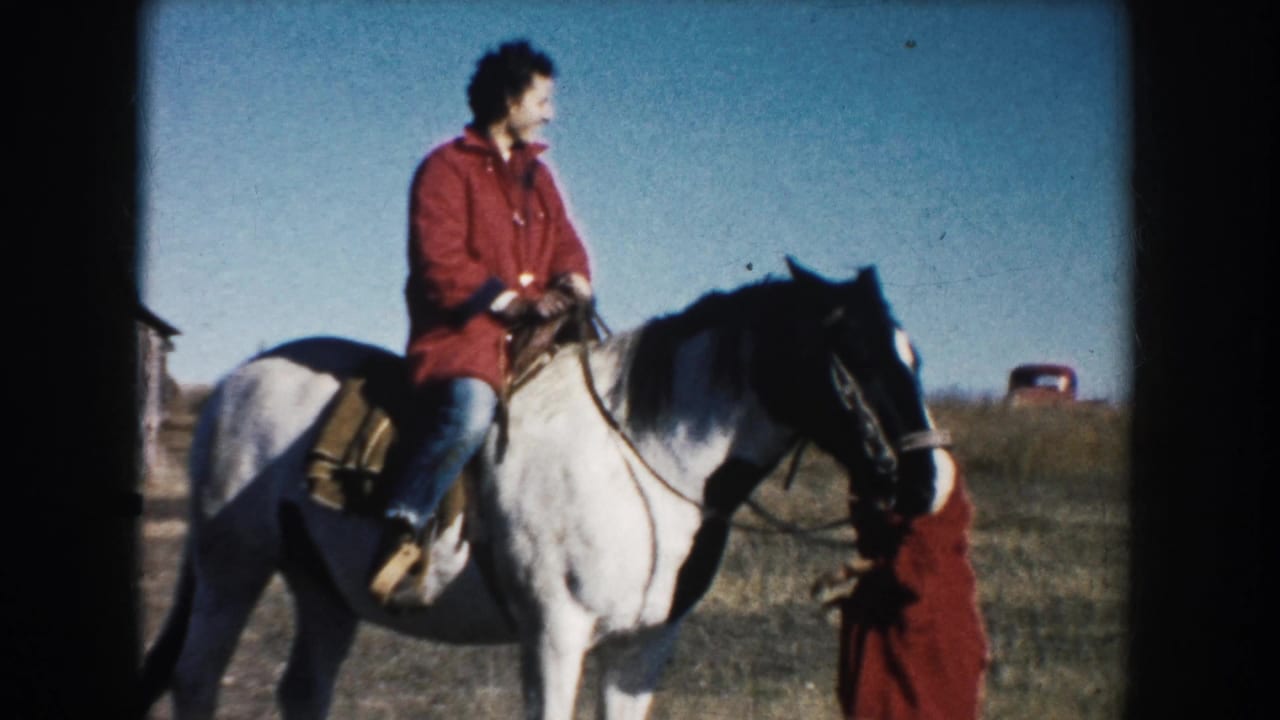 The rider sits upright on a white horse in rural South Dakota while a woman in a red coat stands beside the bridle area.