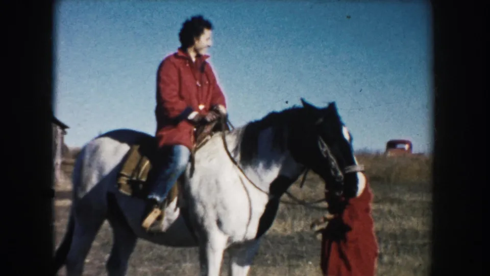 The rider sits upright on a white horse in rural South Dakota while a woman in a red coat stands beside the bridle area.