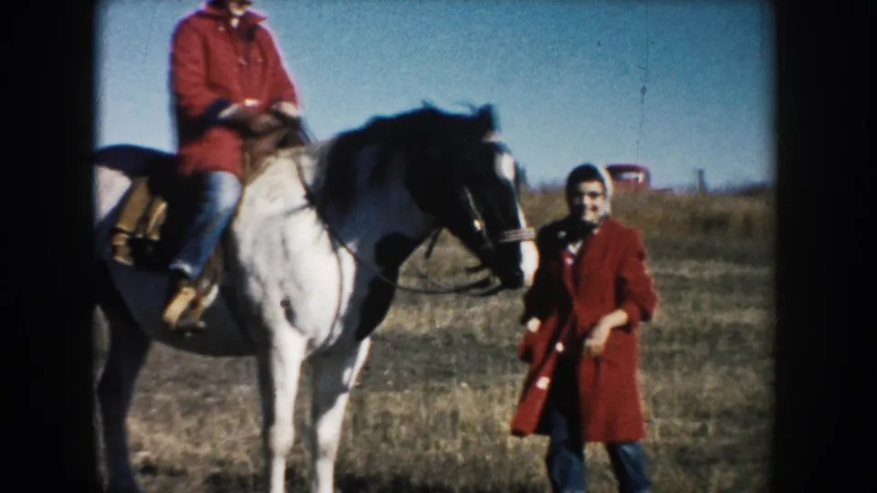 A man remains mounted on the white horse while the woman in red stands nearby, both framed by a grassy South Dakota field.