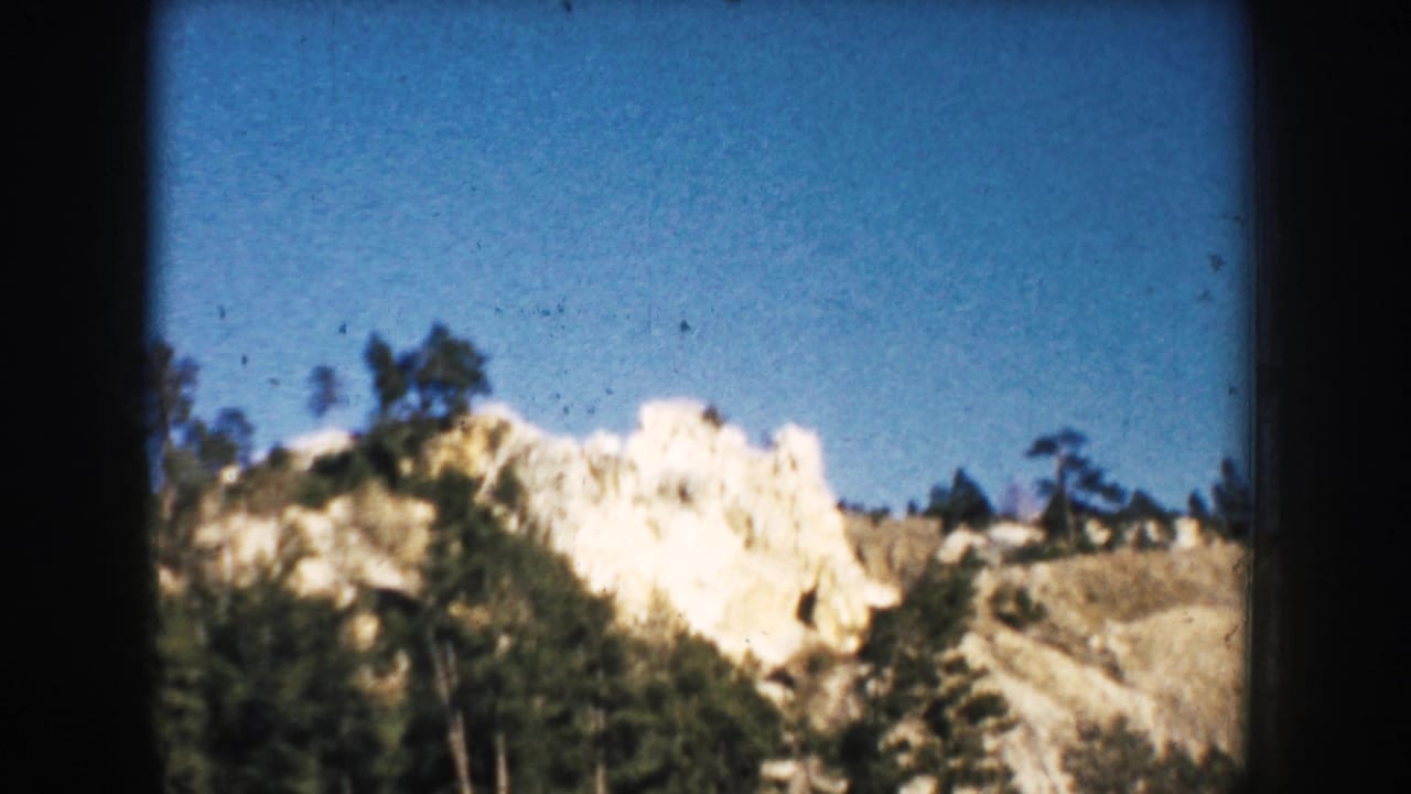 The South Dakota landscape opens to a pale rocky hill or landmark with scattered trees beneath a clear blue sky in 1953 8mm footage.