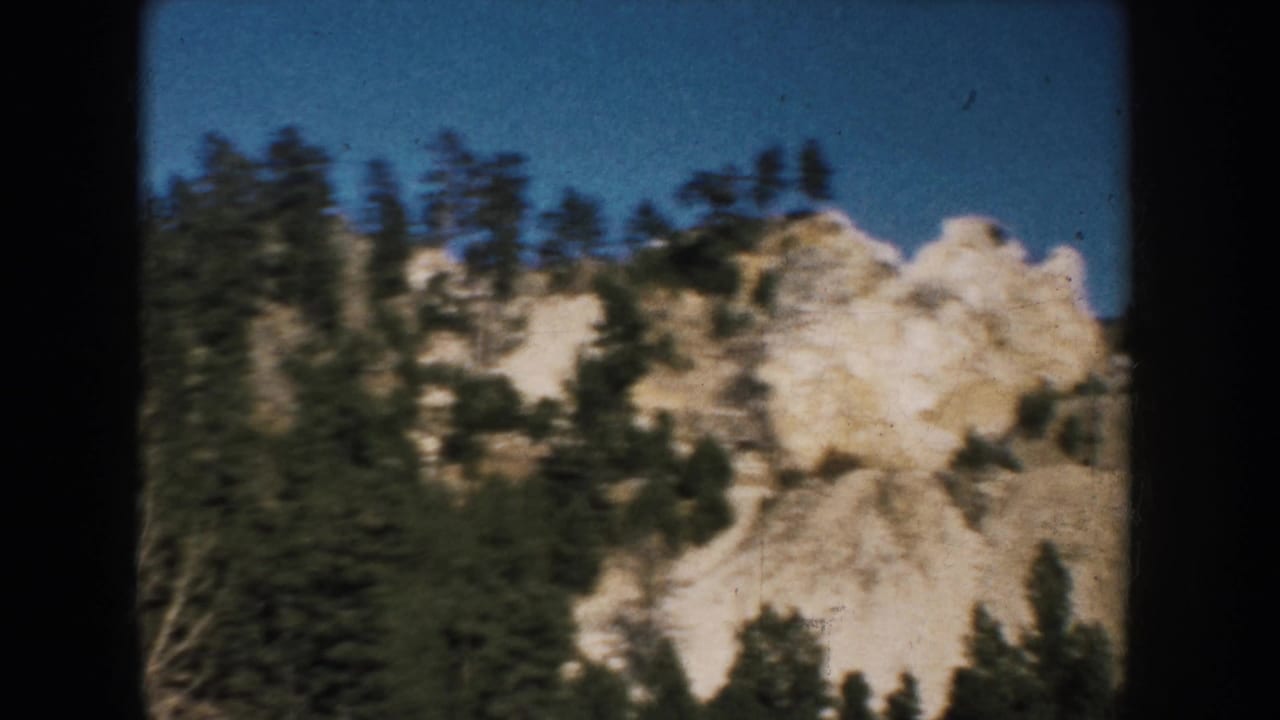 Pine-covered rocky hillside beneath clear blue sky with dark frame borders.