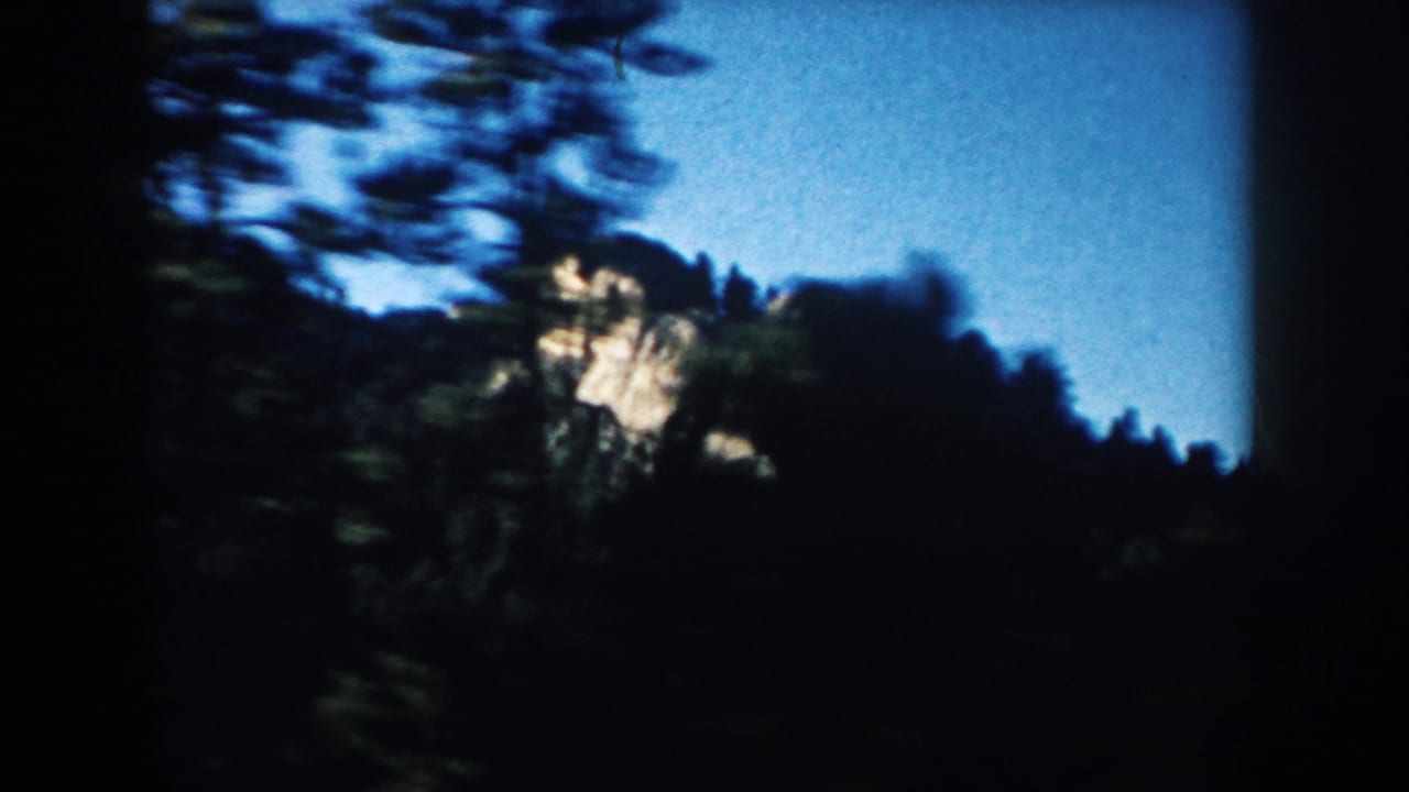 1953 South Dakota 8mm frame with motion-blurred trees and a pale tan rocky slope or mountain face under blue sky.