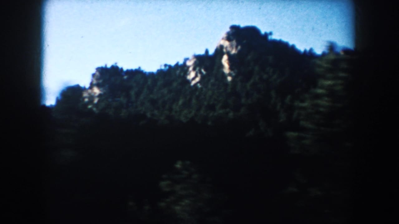South Dakota landscape from the moving car, showing a tree-covered rocky hill under bright blue sky.