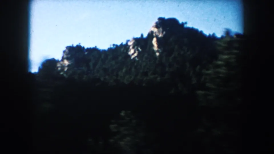 South Dakota landscape from the moving car, showing a tree-covered rocky hill under bright blue sky.