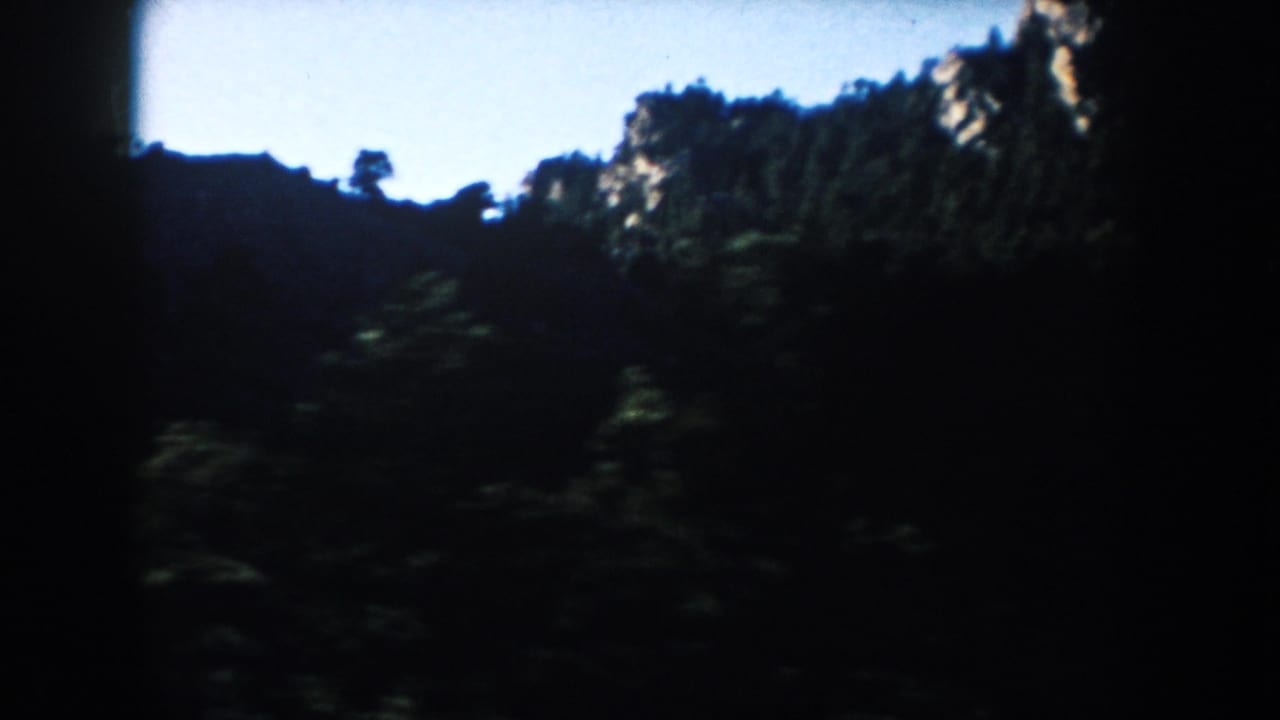 1950s 8mm roadside landscape with shadowed trees and a rocky South Dakota ridge softened by motion.