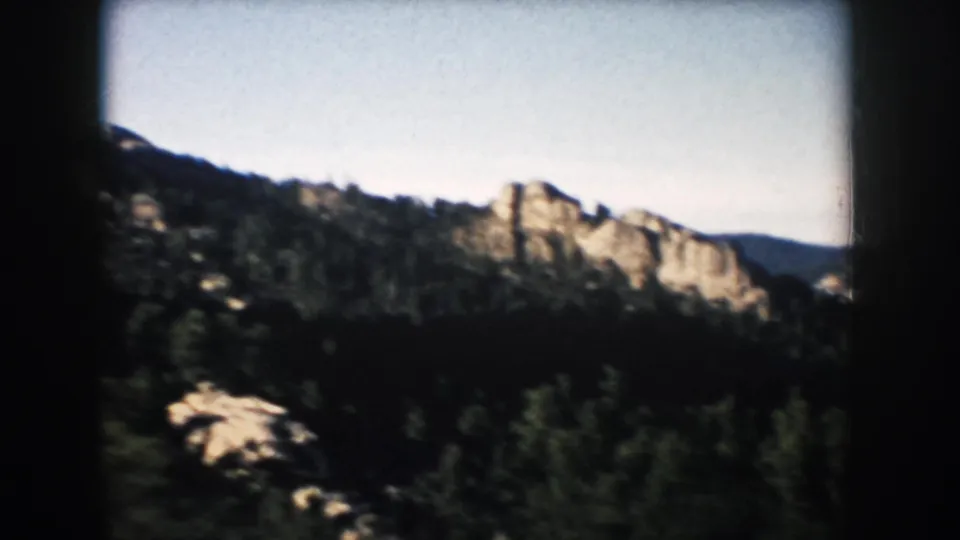 1953 South Dakota 8mm frame showing forested hills and tan rock outcrops under a pale sky.