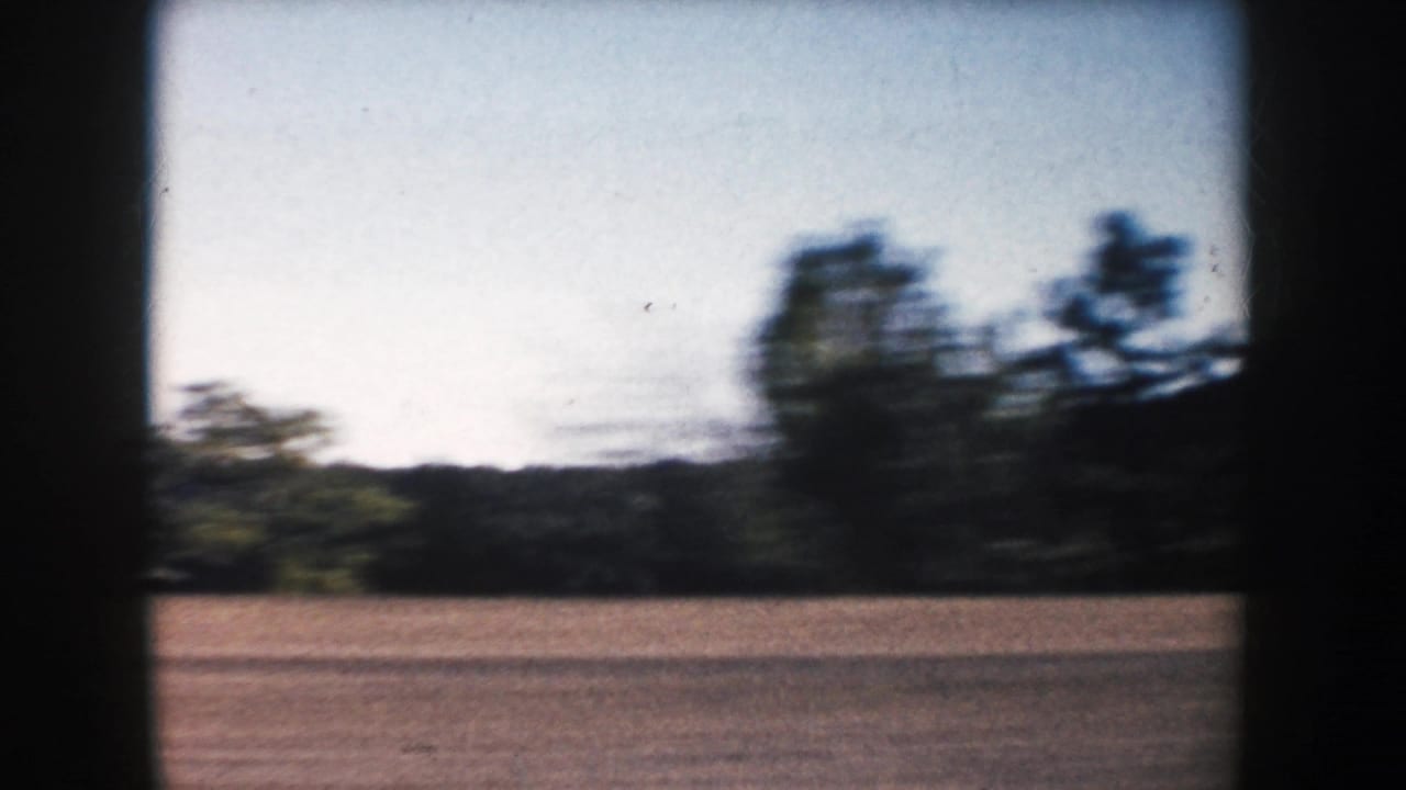 Motion-blurred 1953 South Dakota 8mm frame showing roadside terrain, trees, and distant hills.