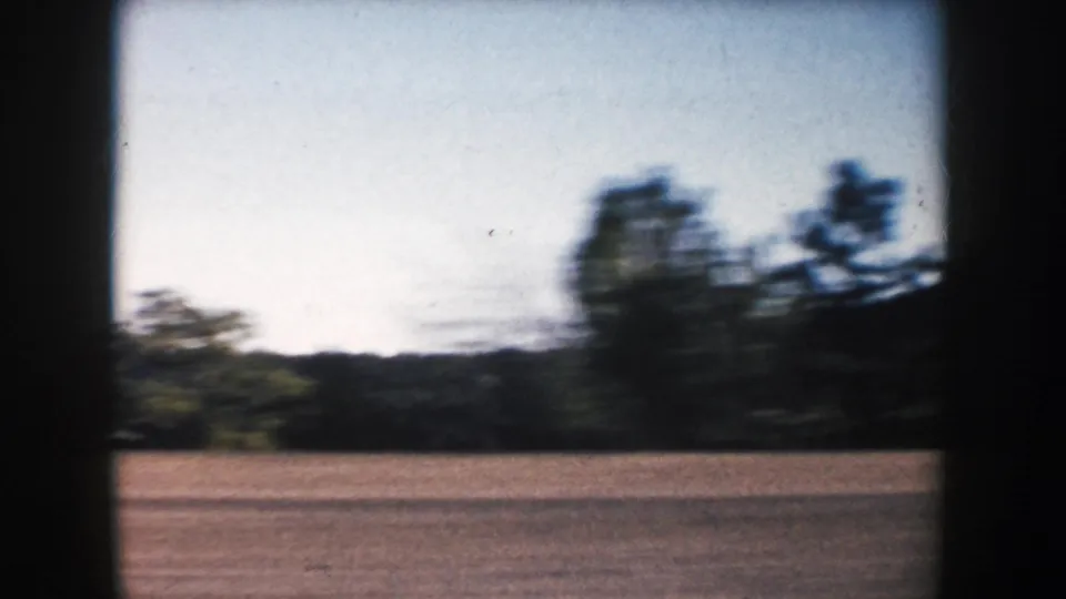 Motion-blurred 1953 South Dakota 8mm frame showing roadside terrain, trees, and distant hills.