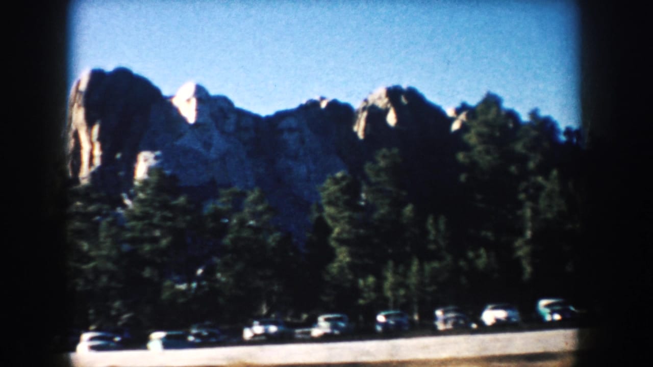 1953 South Dakota 8mm frame of Mount Rushmore viewed past trees and a parking area.