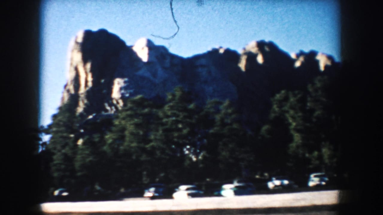 Mount Rushmore above evergreen trees with parked cars in the foreground.