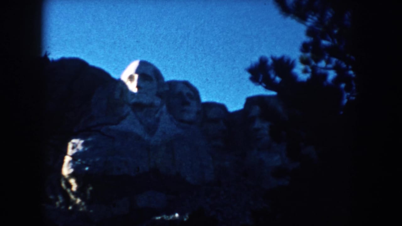 Mount Rushmore appears in a 1953 South Dakota home-movie frame, partly blocked by dark tree silhouettes beneath a clear blue sky.