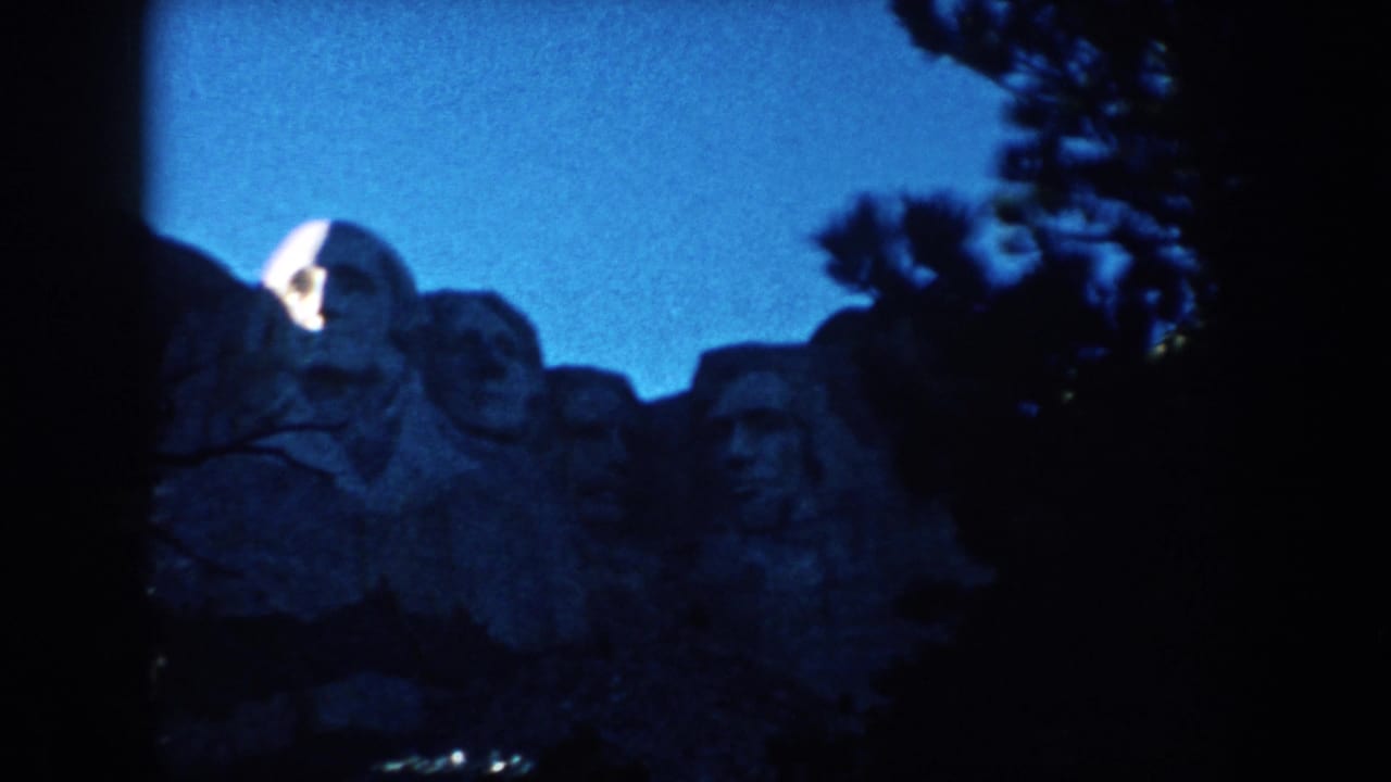1953 8mm view of Mount Rushmore in South Dakota, with the carved faces framed by shadowed trees and saturated blue sky.