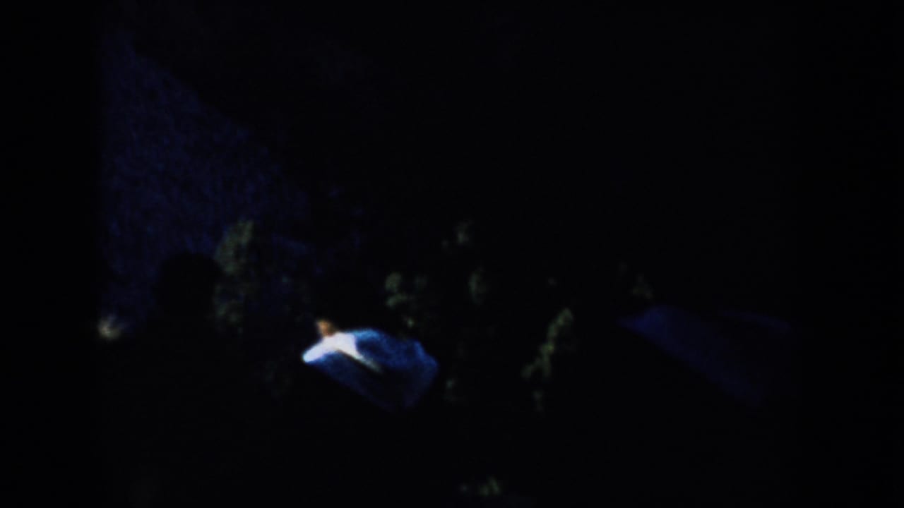 Shadowed 1953 South Dakota home-movie frame showing Mount Rushmore, dark trees, blue sky, and a small distant visitor-like figure below.