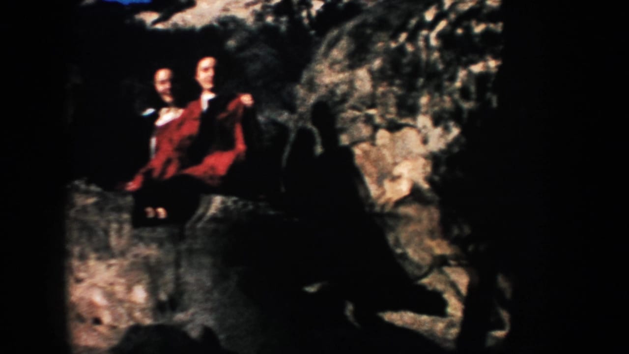 A person in a red coat sits on rocks beside another partially visible figure.