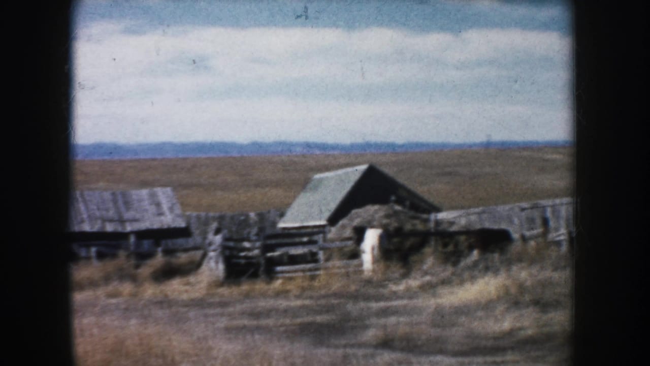 South Dakota farmyard on 8mm film, showing weathered wooden outbuildings, fencing, dry prairie grass, cloudy sky, and distant hills.