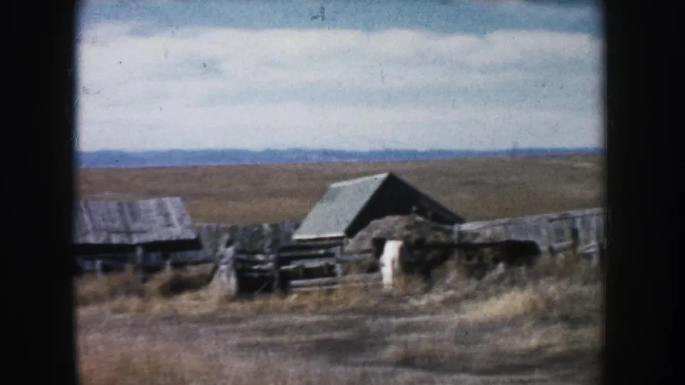 South Dakota farmyard on 8mm film, showing weathered wooden outbuildings, fencing, dry prairie grass, cloudy sky, and distant hills.