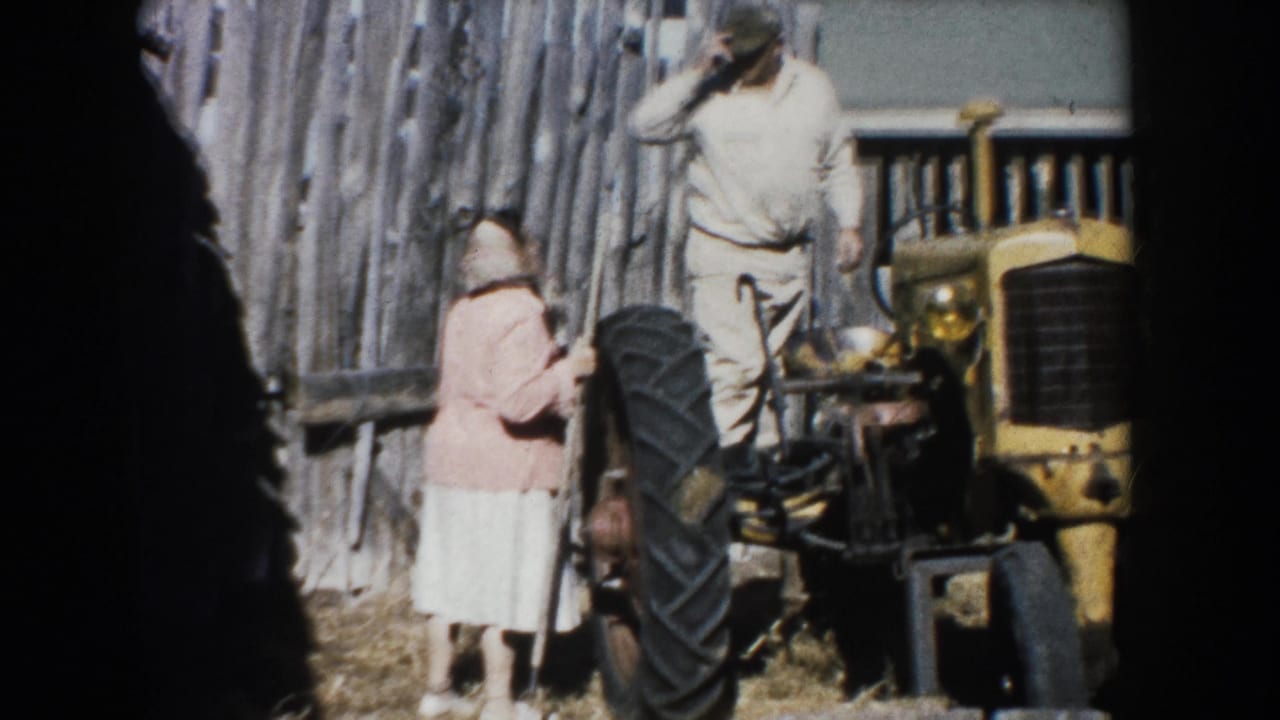 1953 South Dakota farm scene with an adult woman beside a yellow tractor and an adult man standing near a wood fence.