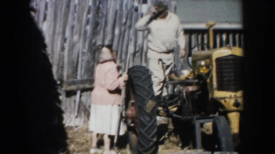 1953 South Dakota farm scene with an adult woman beside a yellow tractor and an adult man standing near a wood fence.