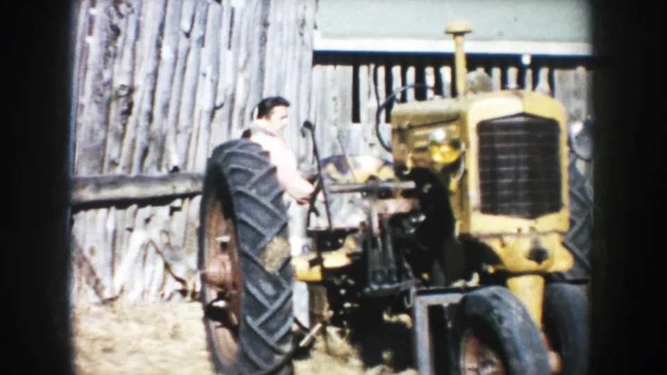 1953 South Dakota farm scene with a man beside a yellow tractor in front of a weathered wooden barn wall.