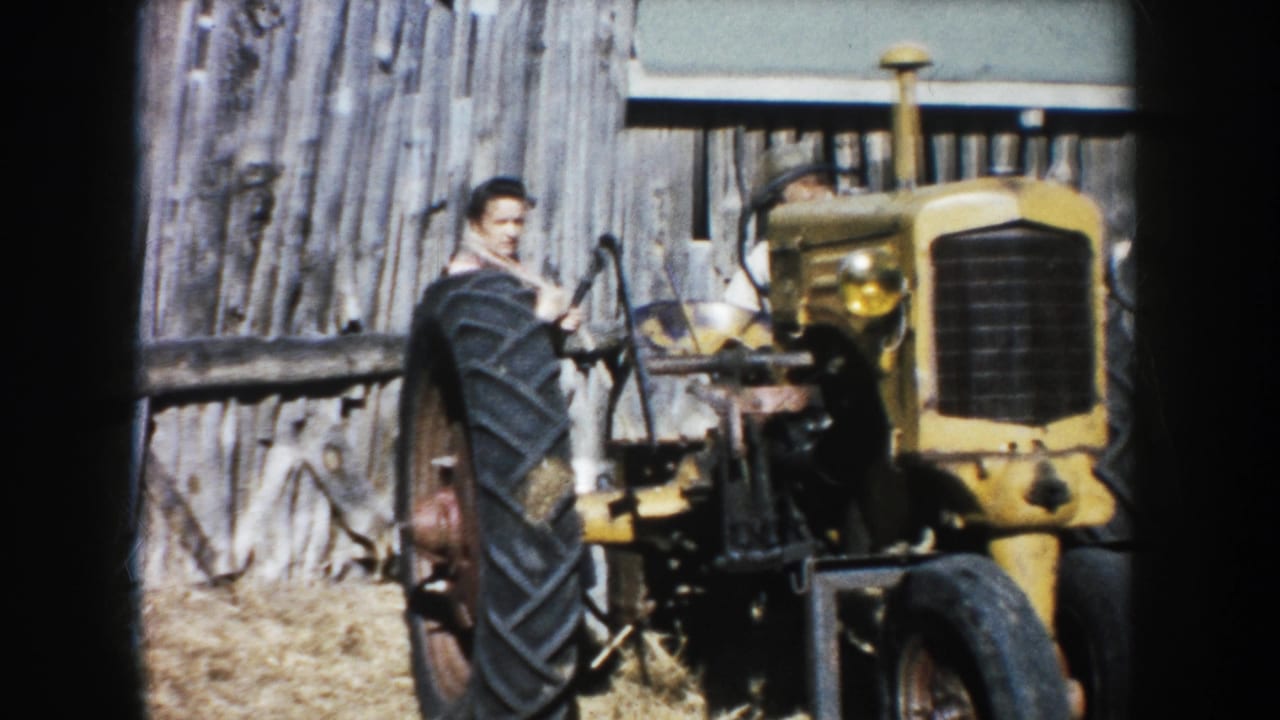 1953 South Dakota 8mm frame showing an adult man at a yellow tractor with barn boards and straw visible around him.
