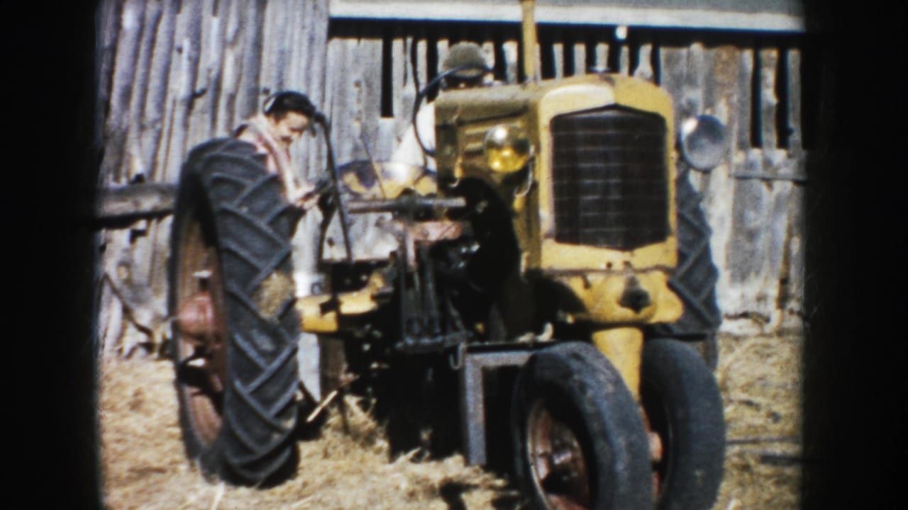 1953 South Dakota farm footage frame with a man beside a yellow tractor, seen close to the barn wall and dry ground.