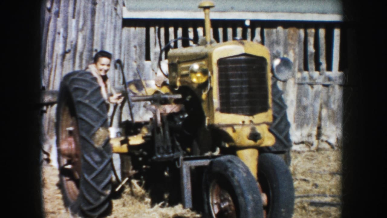 Man standing beside a yellow tractor in a farmyard near wooden fencing.