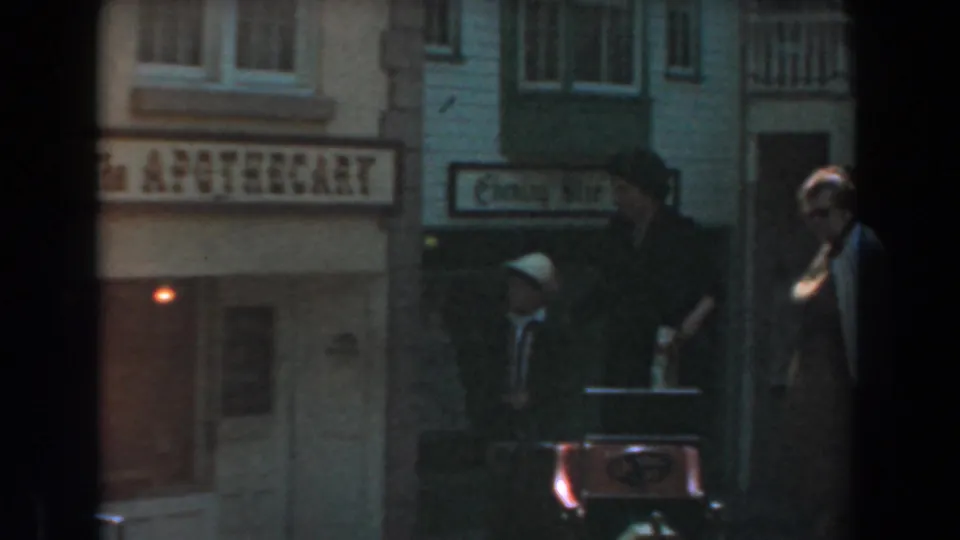 A 1961 home-movie frame at Knott's Berry Farm shows adults near the apothecary sign, with a woman in a light hat beside a red stroller or cart.