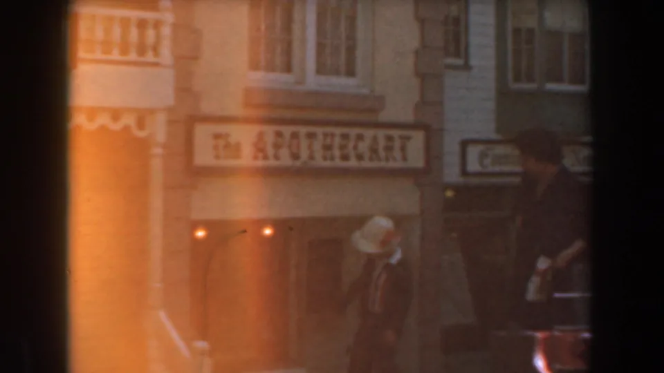The 1961 Knott's Berry Farm street view is partly veiled by orange 8mm flare as visitors pass the apothecary storefront and nearby signage.