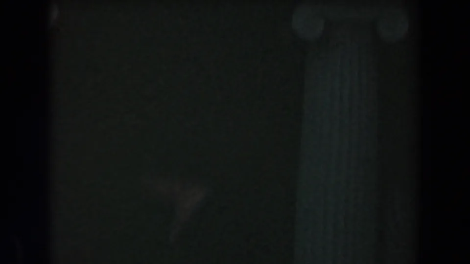Dim 1961 home-movie frame with partial columns, dark architectural detail, and a possible adult figure at the right edge.