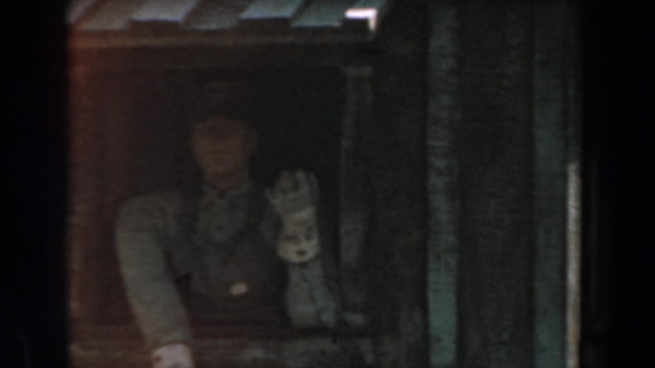 Person waving beside dark station piping and a red rail car area.