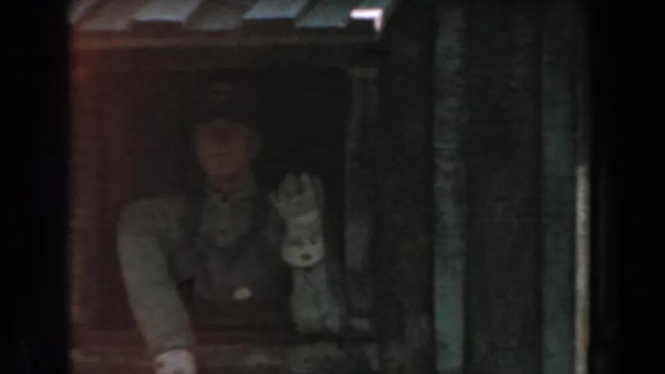 At Knott's Berry Farm in 1961, a person appears beside dark piping and woodwork, raising a hand toward the camera near a rail car setting.