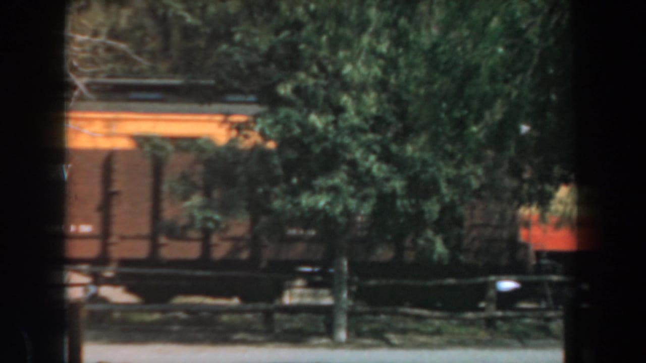 Brown and yellow train cars pass behind trees and a fence, seen through dark window-frame borders.